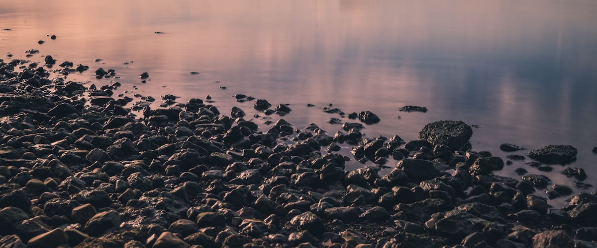 A calm lake with a rocky shoreline and a bridge in the distance.