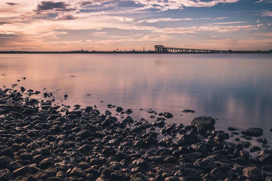 A calm lake with a rocky shoreline and a bridge in the distance.