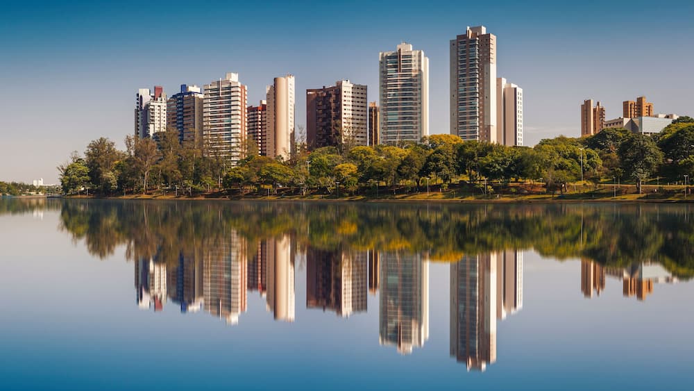 A city skyline reflected in a calm lake with trees and greenery along the shore.