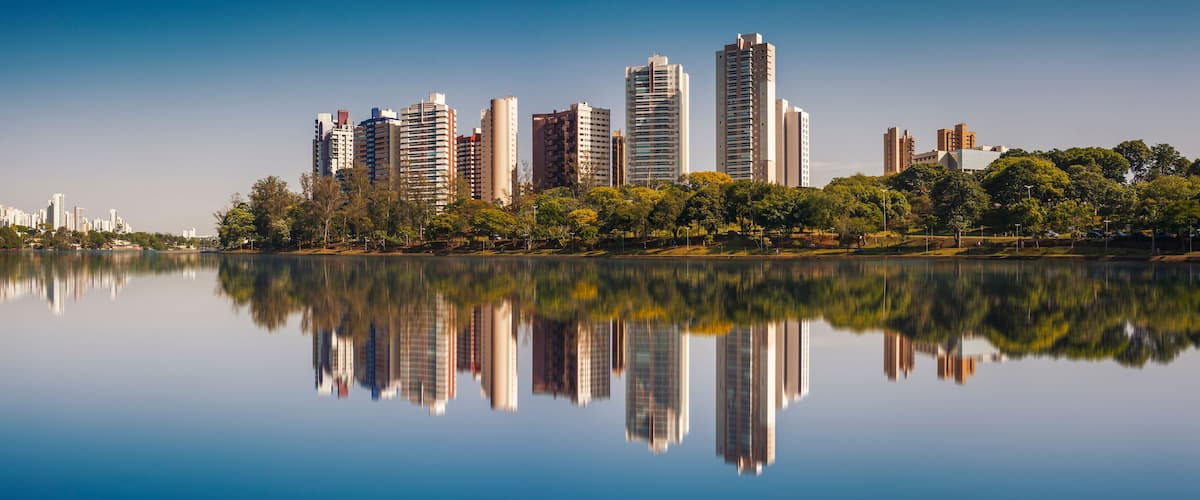 A city skyline reflected in a calm lake with trees and greenery along the shore.