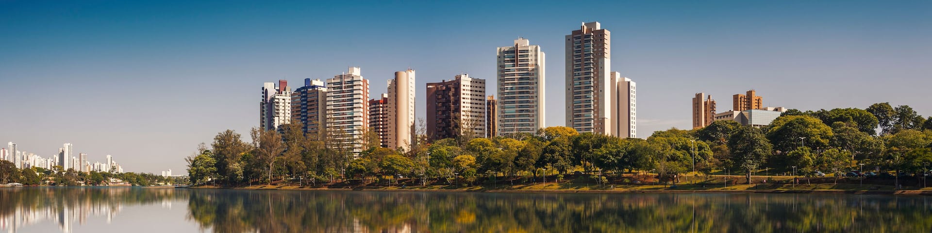 A city skyline reflected in a calm lake with trees and greenery along the shore.