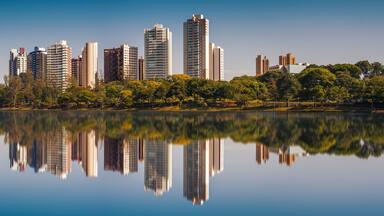 Un horizonte urbano reflejado en un lago tranquilo con árboles y vegetación a lo largo de la orilla.
