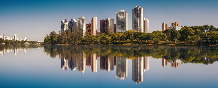 Linha do horizonte urbana refletida em um lago calmo, com árvores e vegetação ao longo da margem.