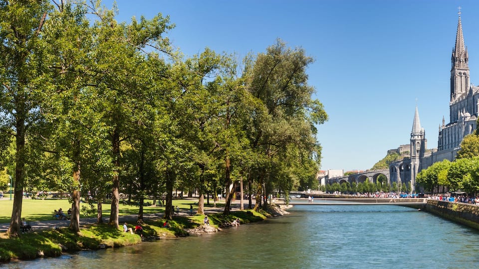 A river with a stone bridge, trees, and a historic building with a tall spire.