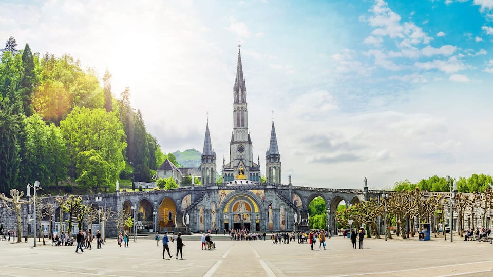A historic church with a tall spire, surrounded by trees and a large open plaza with people walking.