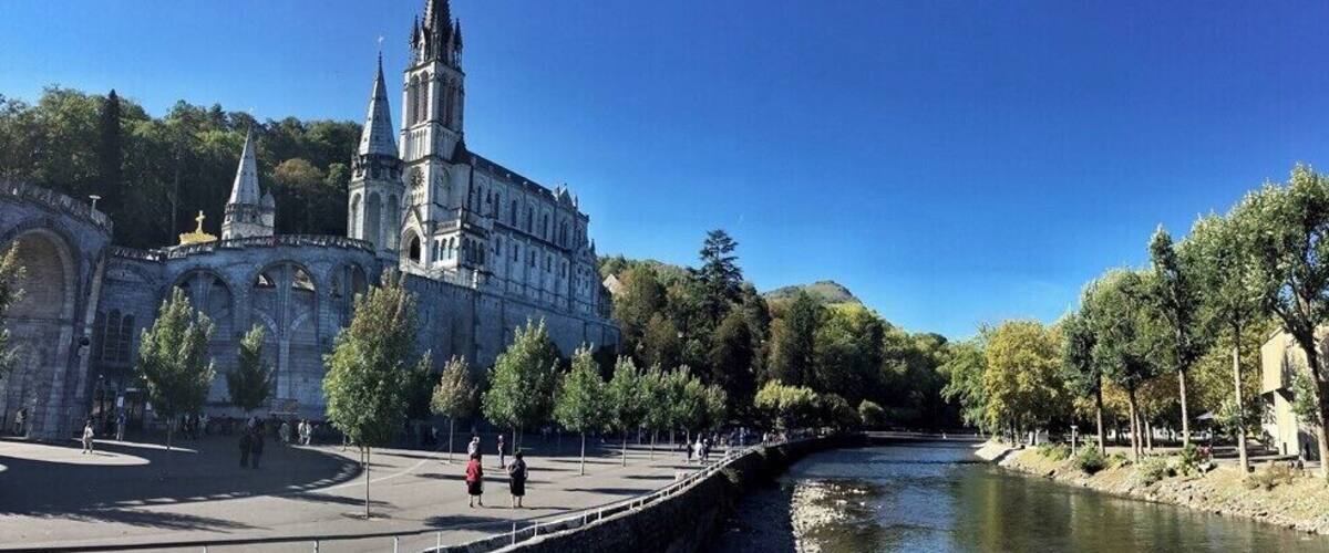 A historic building with a tall spire, a river, and a tree-lined promenade.