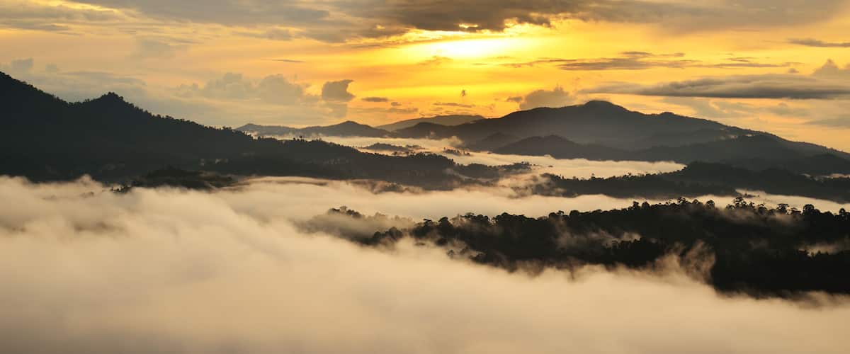 A mountainous landscape with a sunrise or sunset, clouds, and a layer of fog or mist covering the lower elevations.