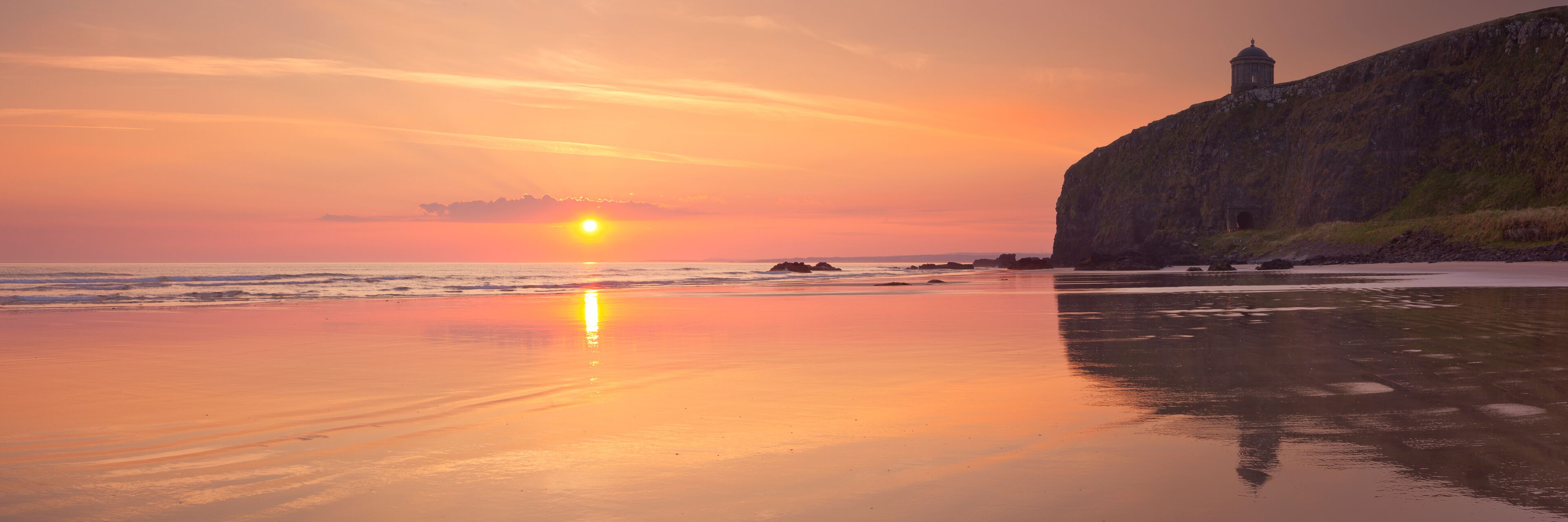 Atardecer sobre una playa con un acantilado rocoso y un faro.