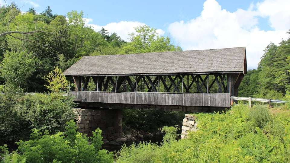 A wooden covered bridge over a rocky stream, surrounded by lush greenery and trees.