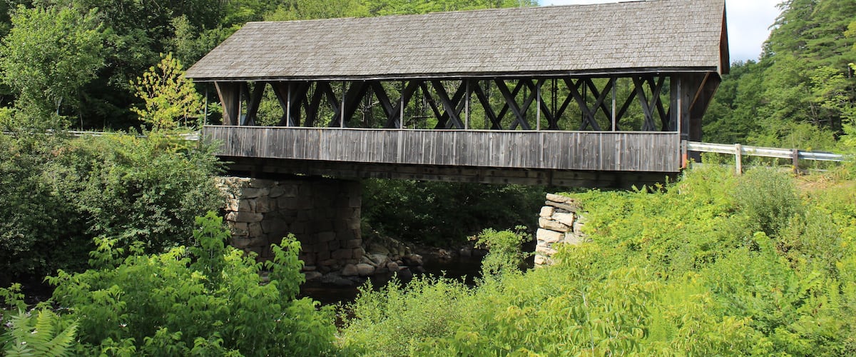 A wooden covered bridge over a rocky stream, surrounded by lush greenery and trees.