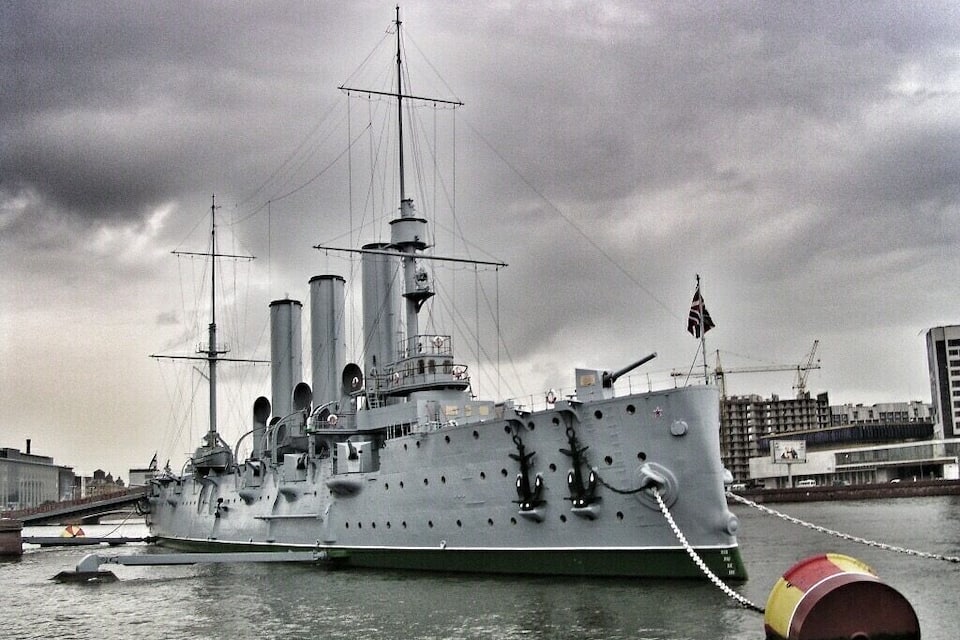 Ein großes Kriegsschiff liegt im Hafen, im Hintergrund ein bewölkter Himmel.