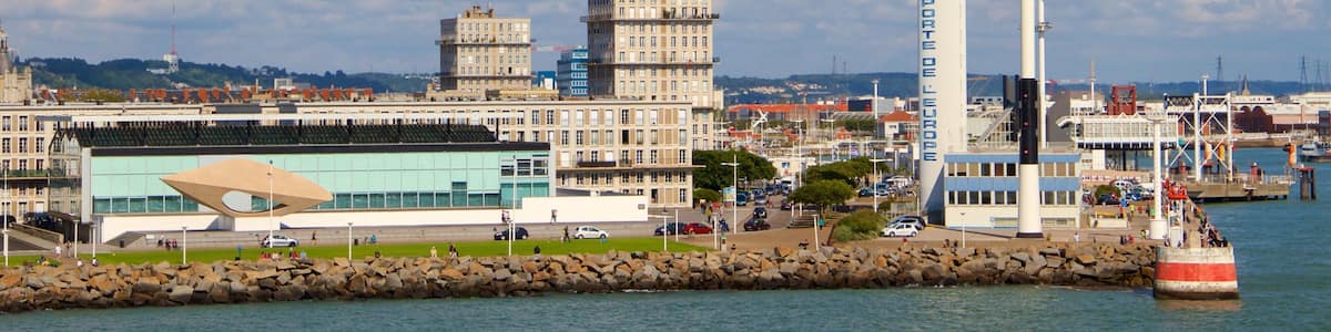 Un paysage urbain côtier avec un phare imposant, un bâtiment moderne doté d’un toit original et une tour historique.