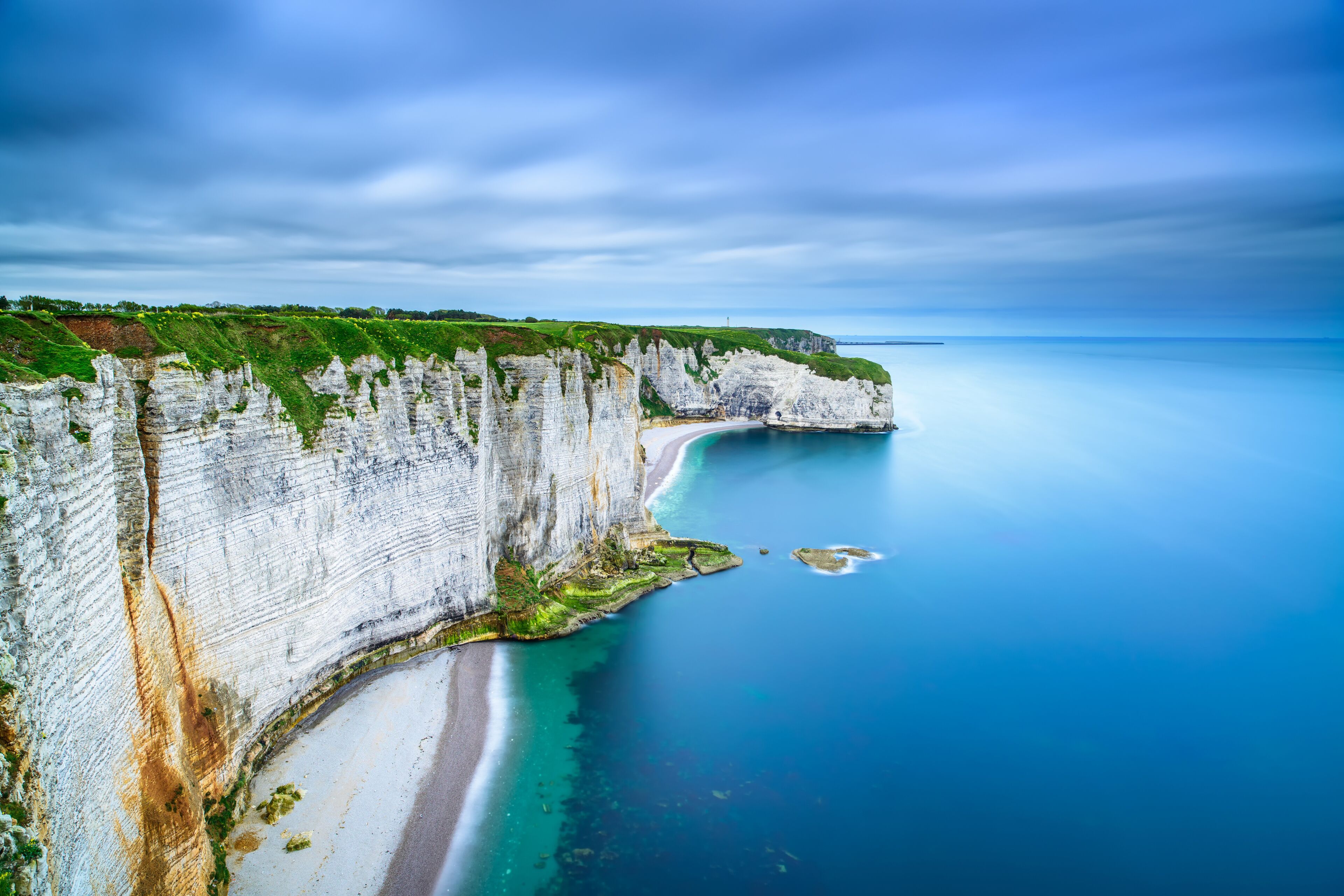 A coastal cliff with white stratified rock layers, a sandy beach, and a calm blue sea.