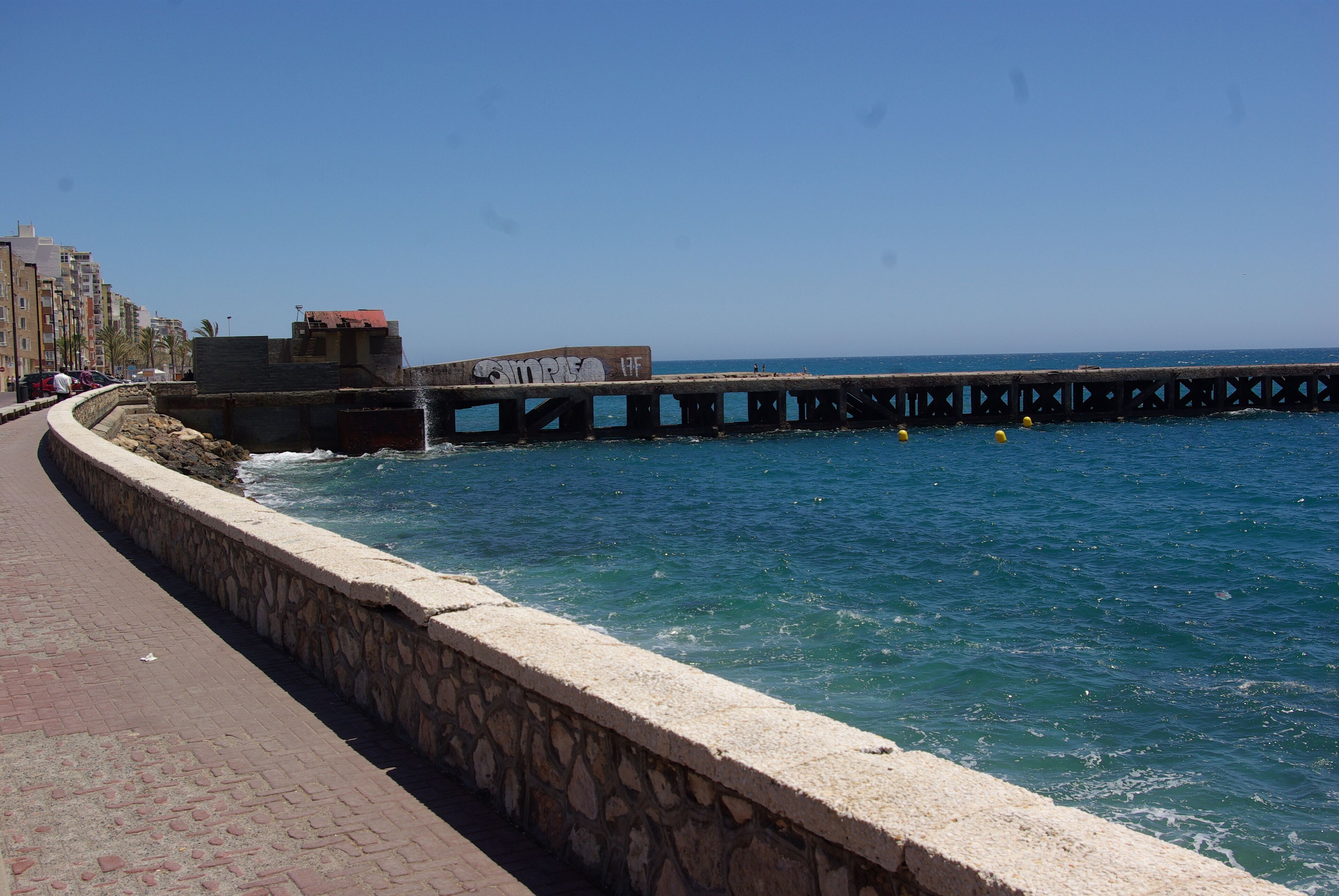 A coastal walkway with a stone wall, a pier extending into the sea, and buildings in the background.
