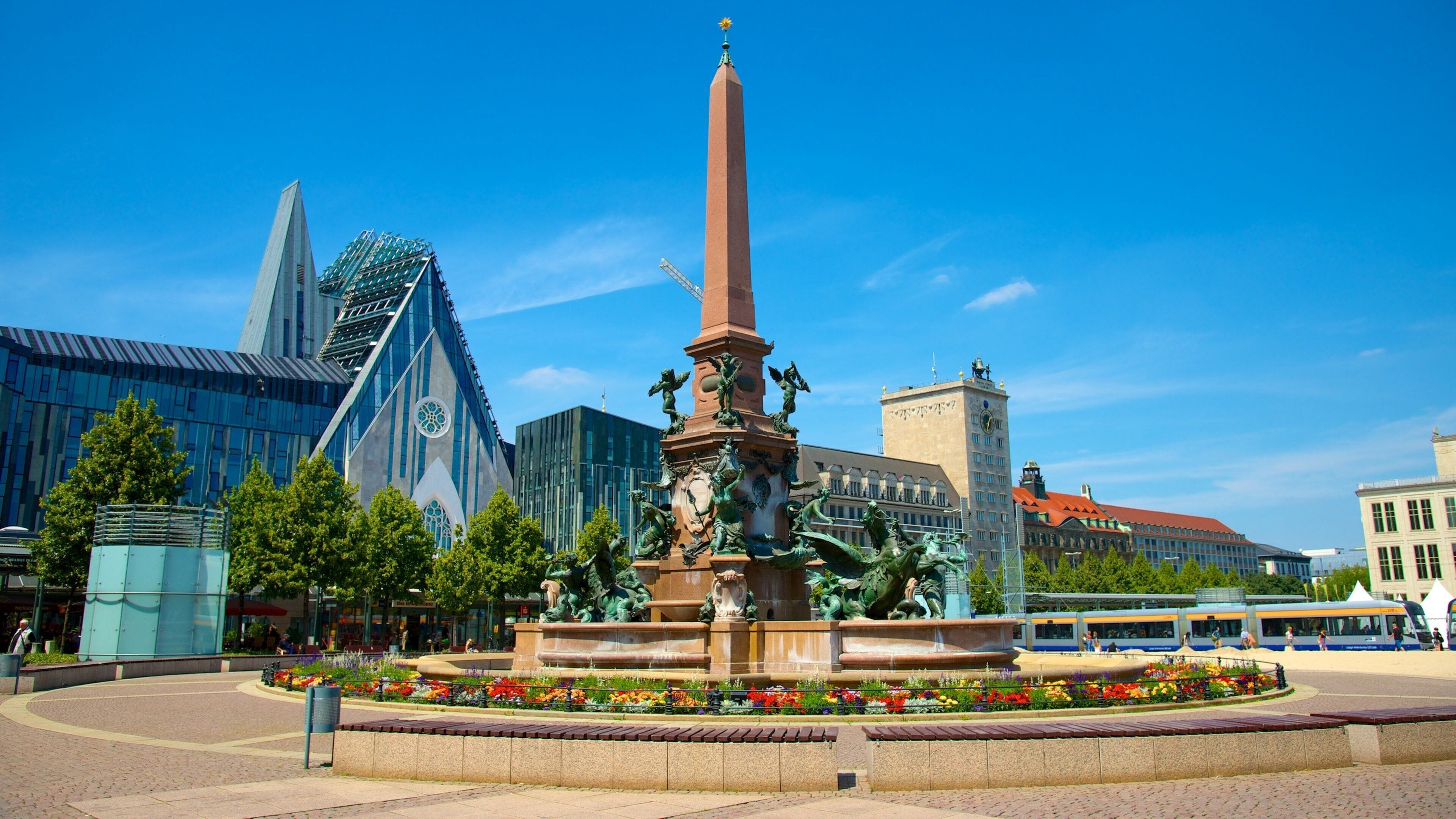 A monument with statues, surrounded by a flower garden and modern buildings.