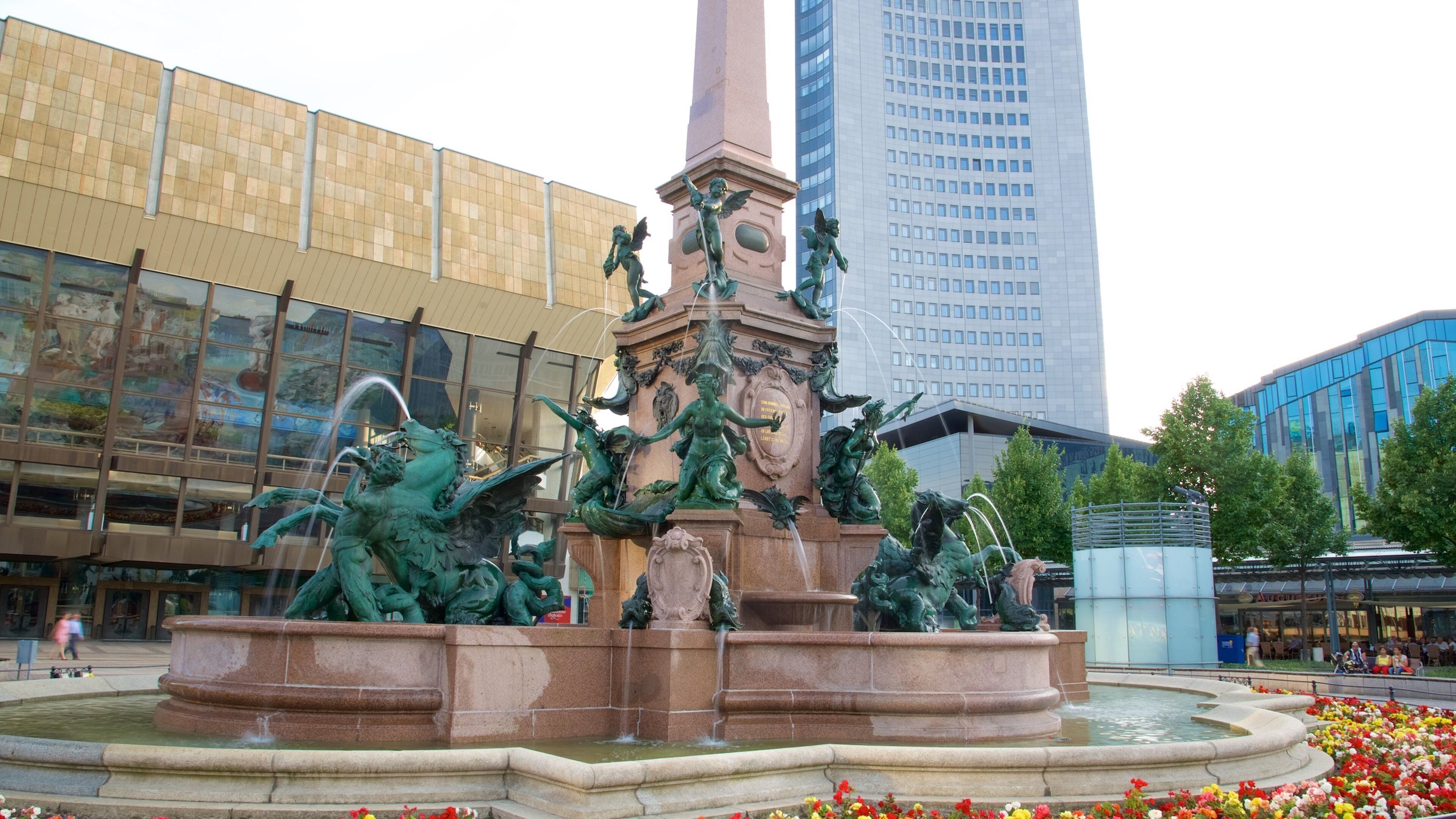 A classical fountain with statues in a plaza, surrounded by flowers and modern buildings.