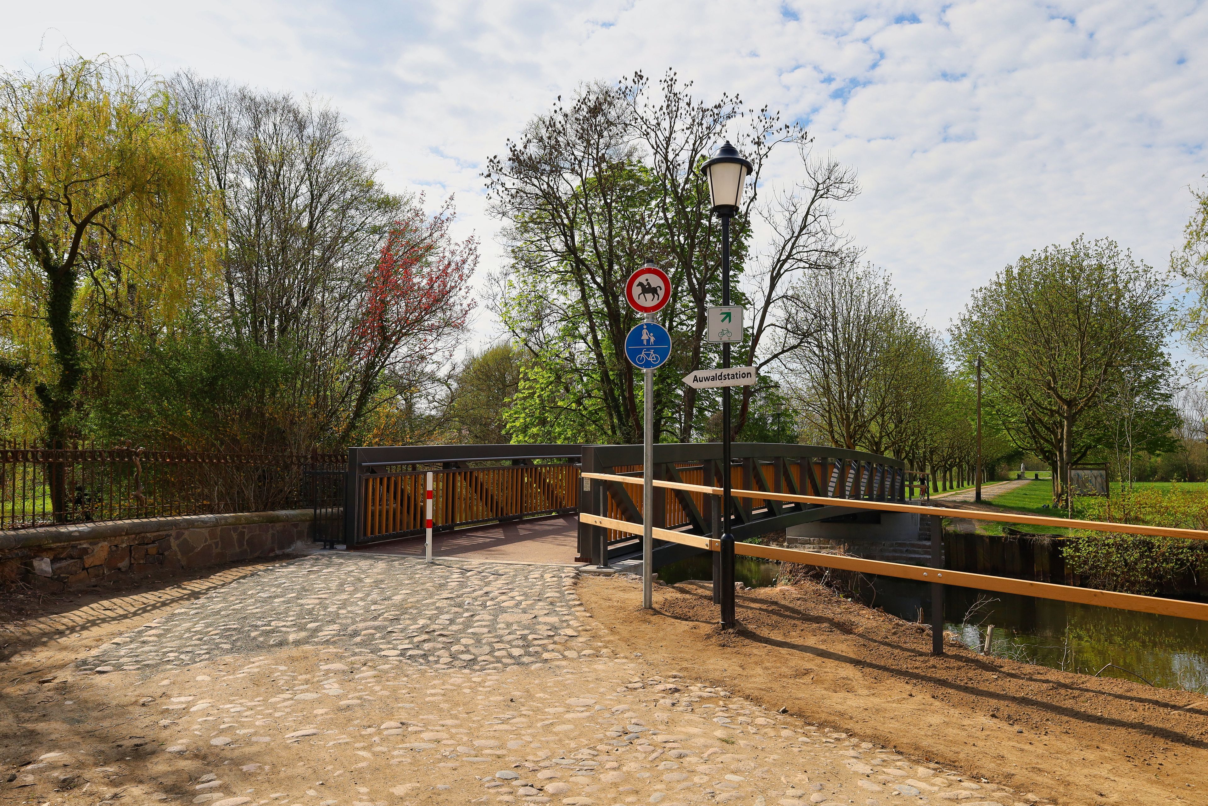 Un sendero en un parque con un puente de madera, un área pavimentada en piedra y un letrero que indica zona de paso prohibido para caballos.