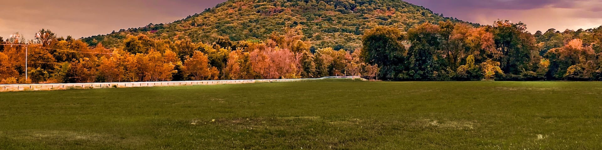 A grassy field with a hill covered in autumn-colored trees and a dramatic sky.