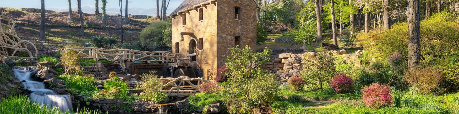 A stone mill house, a wooden bridge, and a flowing stream in a lush garden.