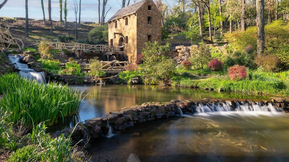A stone mill house, a wooden bridge, and a flowing stream in a lush garden.