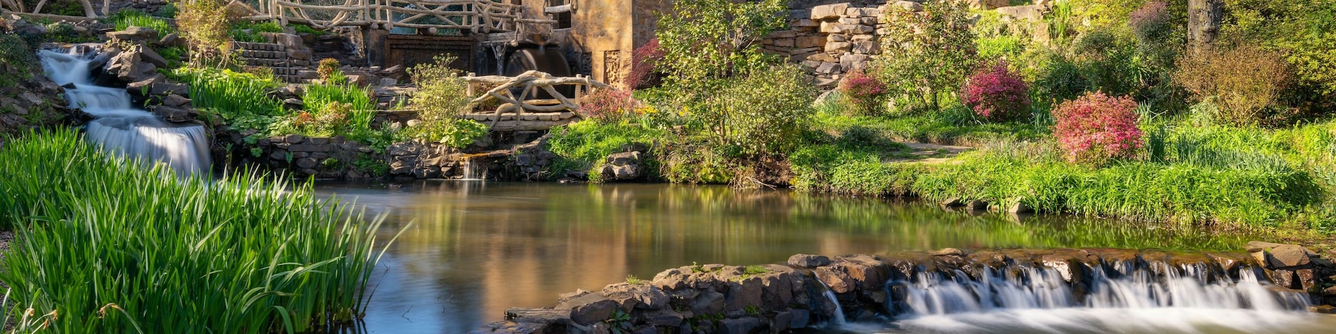 A stone mill house, a wooden bridge, and a flowing stream in a lush garden.