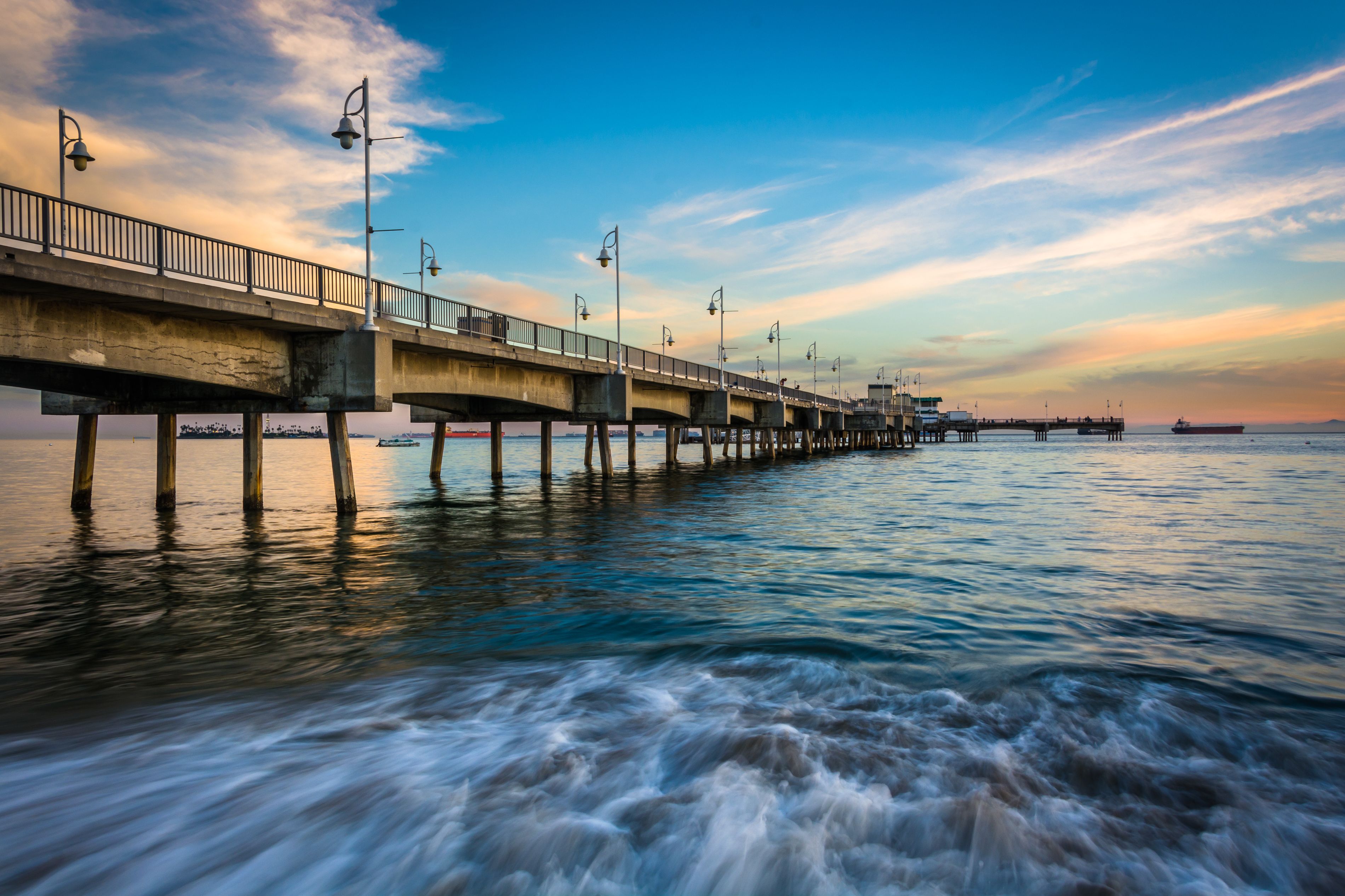 A long pier extending into the water with street lamps and a sunset in the background.