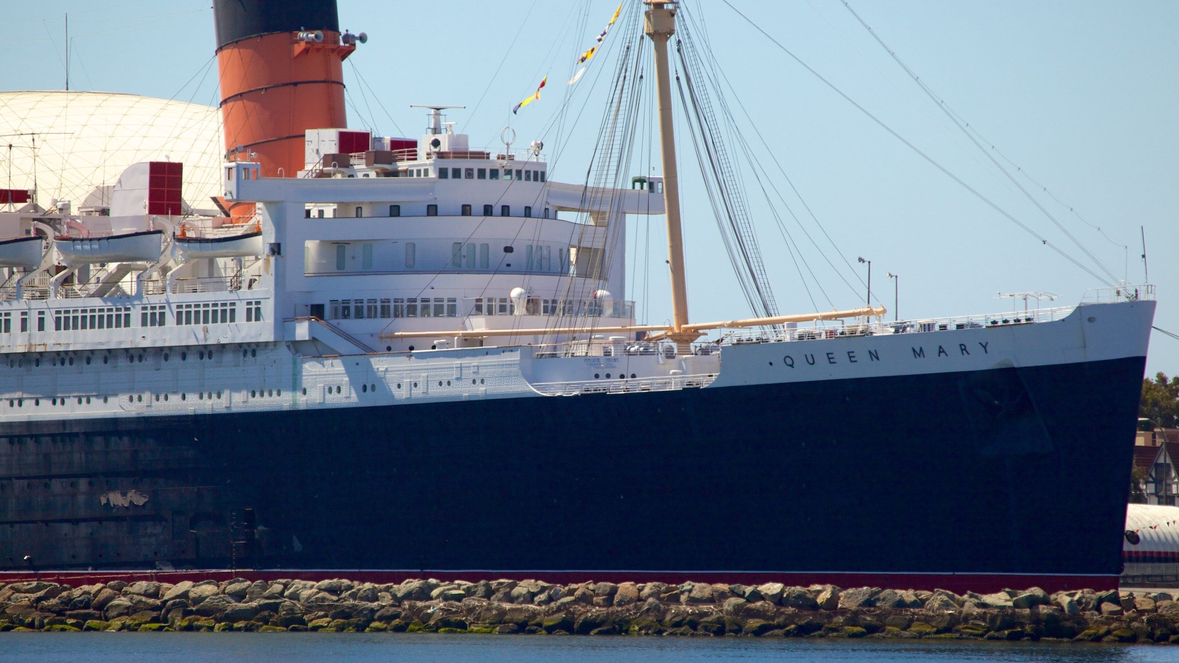 Un gran barco llamado 'Queen Mary' atracado en un puerto con una grúa y una estructura en forma de cúpula al fondo.