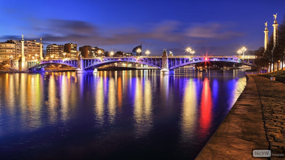 A brightly lit bridge at night with reflections on the water, a cityscape in the background, and a statue on the right.