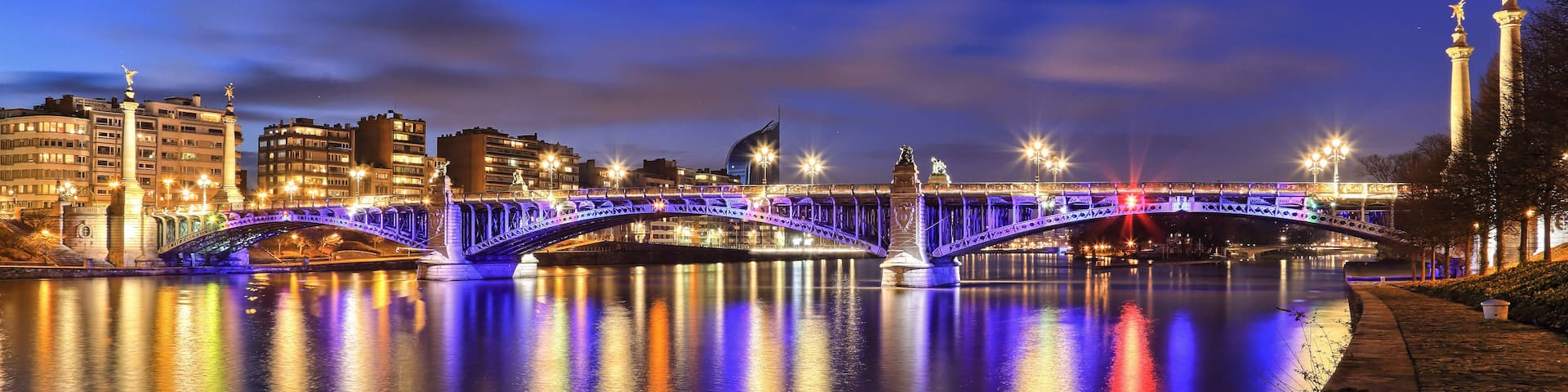 A brightly lit bridge at night with reflections on the water, a cityscape in the background, and a statue on the right.