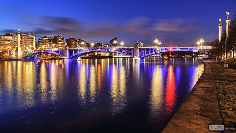 A brightly lit bridge at night with reflections on the water, a cityscape in the background, and a statue on the right.