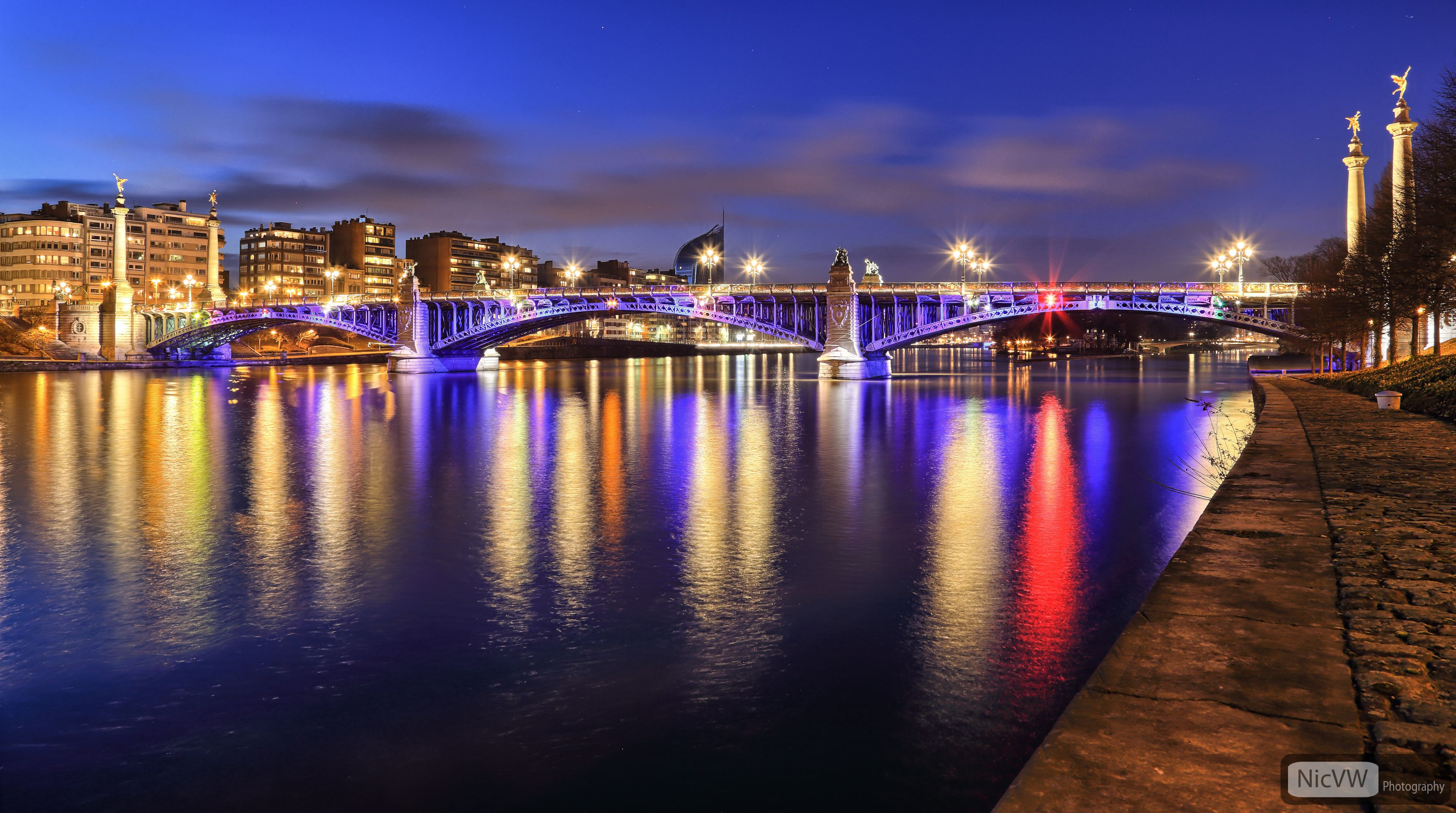 A brightly lit bridge at night with reflections on the water, a cityscape in the background, and a statue on the right.