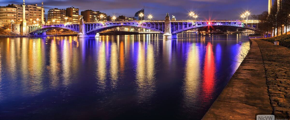 A brightly lit bridge at night with reflections on the water, a cityscape in the background, and a statue on the right.