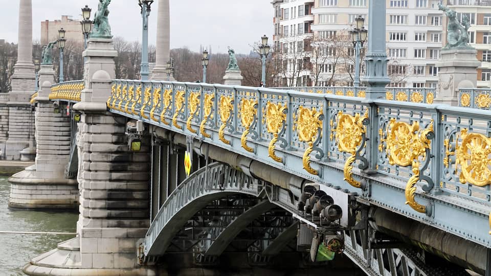 A bridge with ornate golden decorations and statues, spanning over a body of water with buildings in the background.