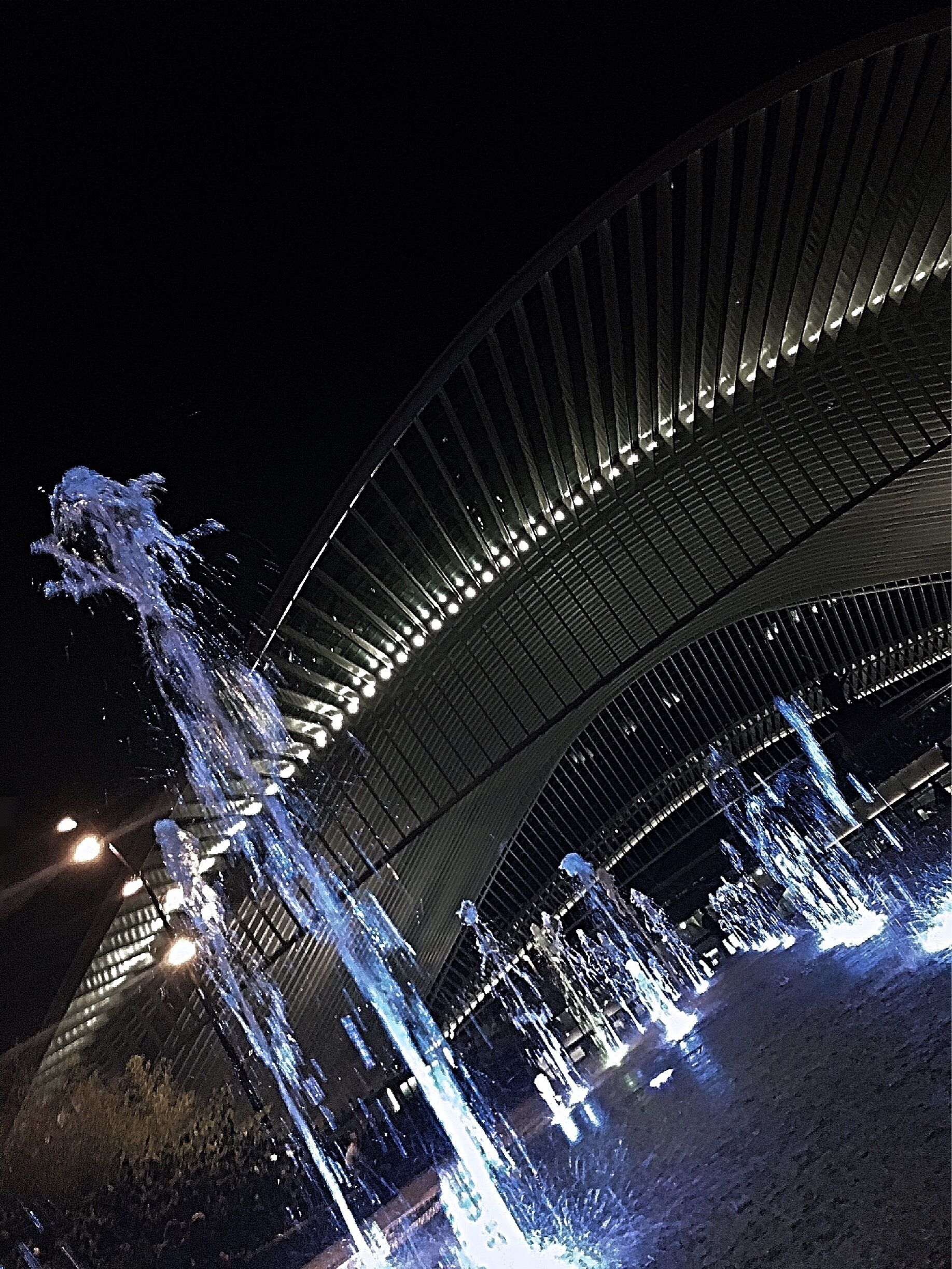 Vista noturna de uma grande ponte iluminada, com fontes d'água caindo pelas laterais.