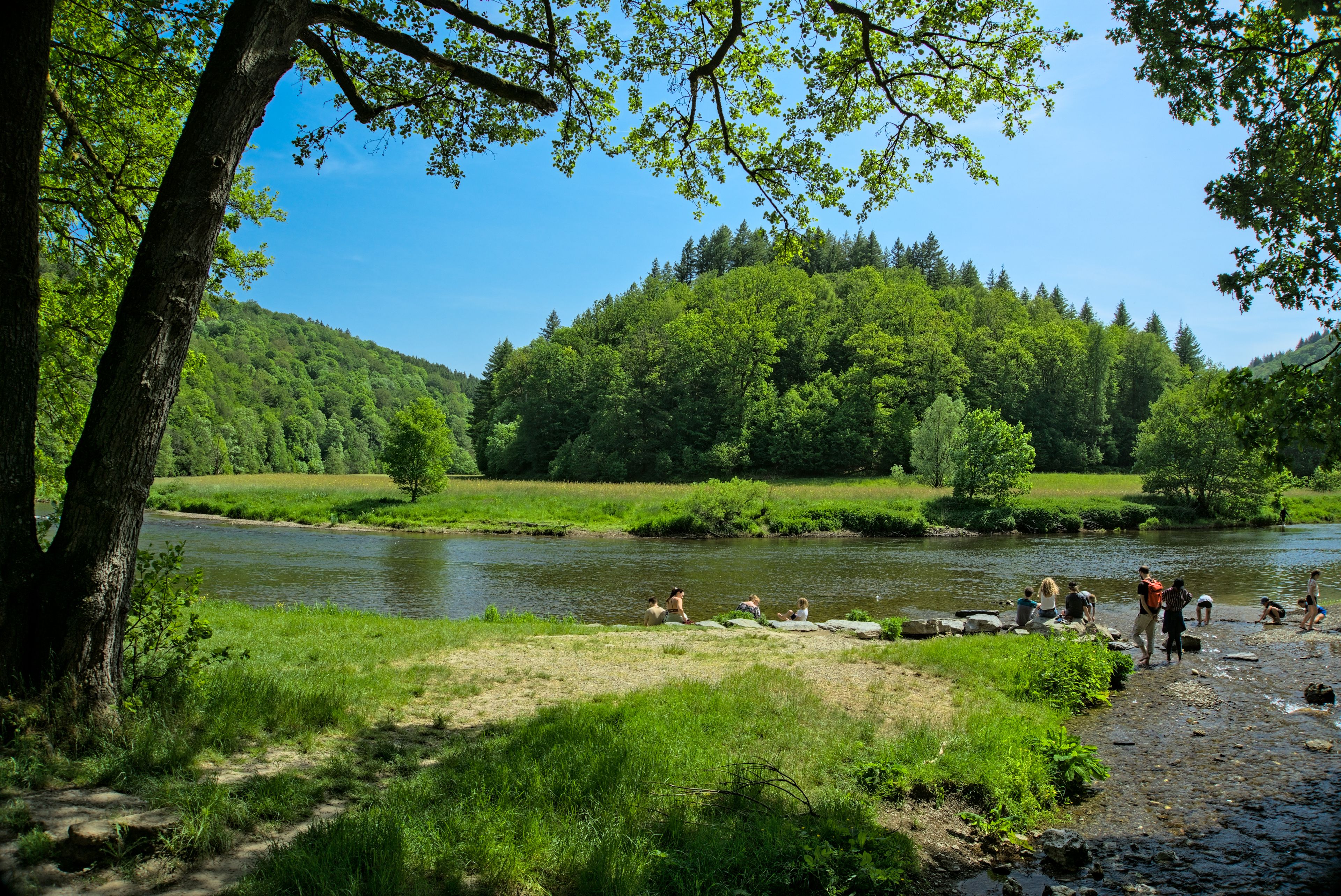 A river with people near the shore, surrounded by lush greenery and hills.