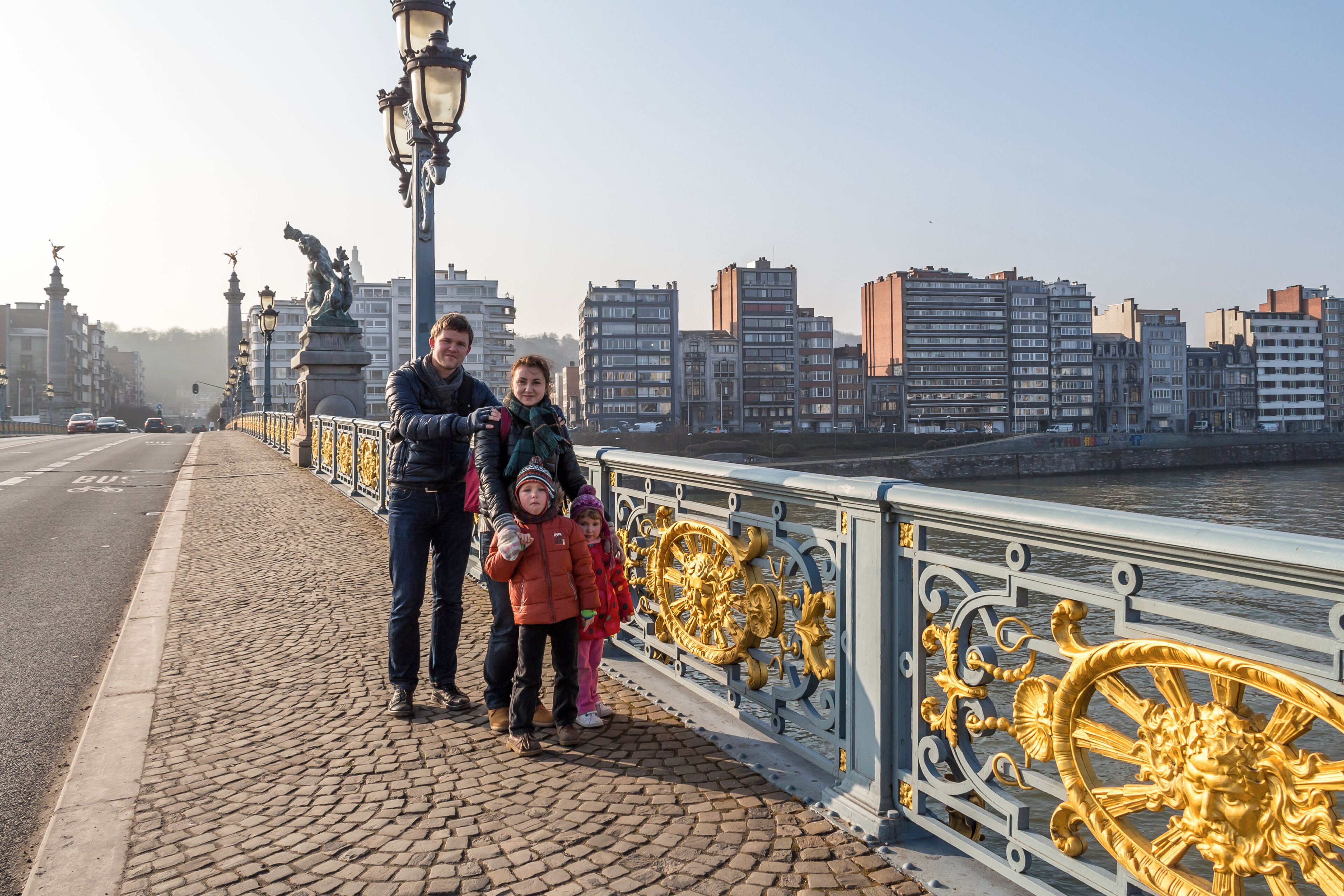 A cobblestone bridge with ornate golden railings, a river, and modern buildings in the background.