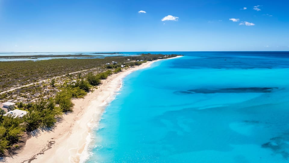 A coastal landscape with a sandy beach, clear turquoise waters, and dense vegetation.