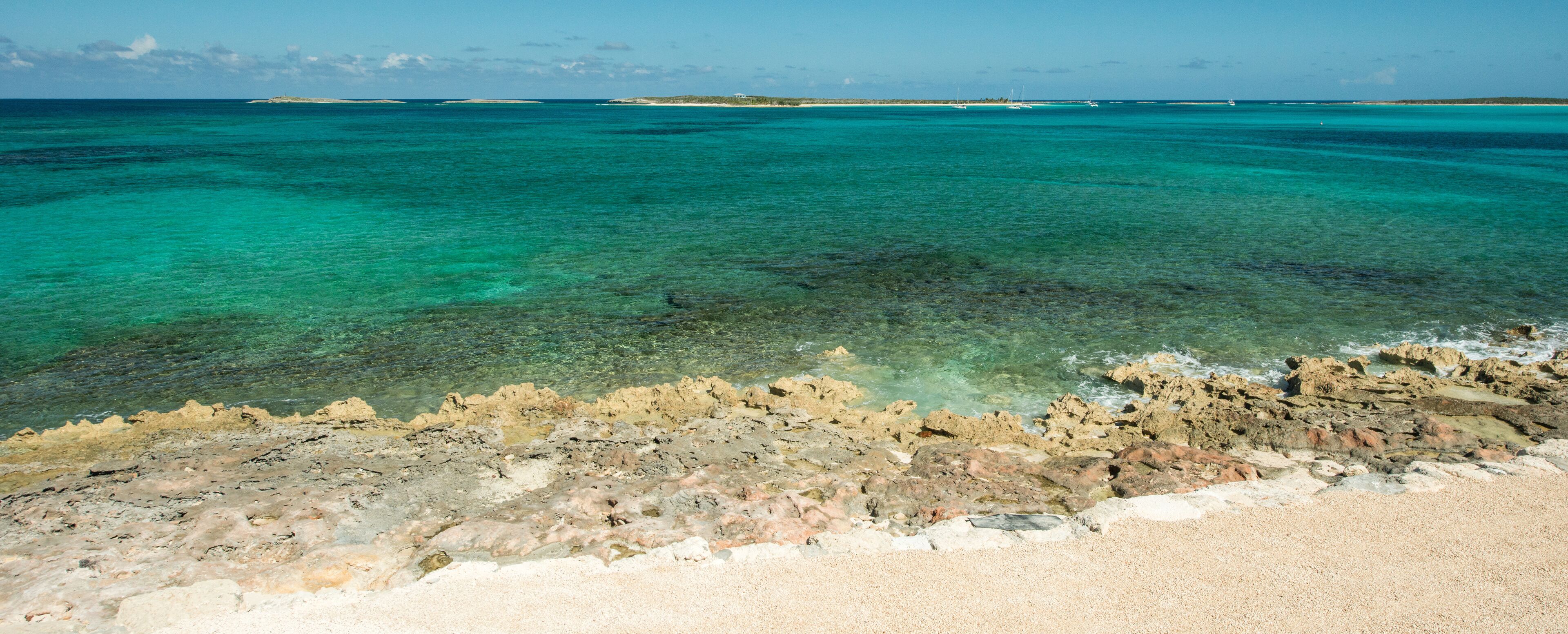 A coastal landscape with clear turquoise waters, rocky shoreline, and distant islands.