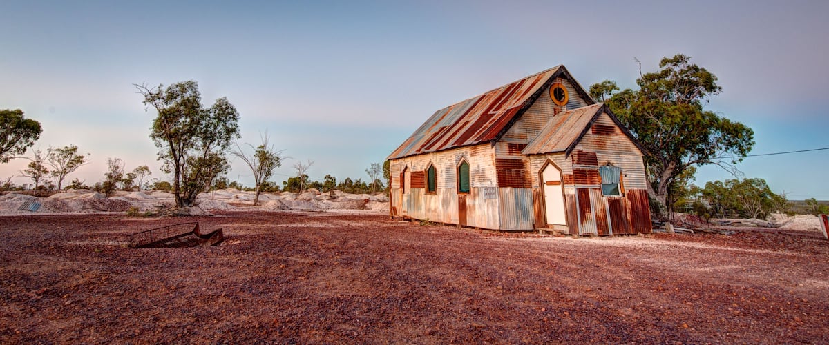 An old, weathered church with a corrugated metal roof, surrounded by sparse vegetation and rocky terrain.