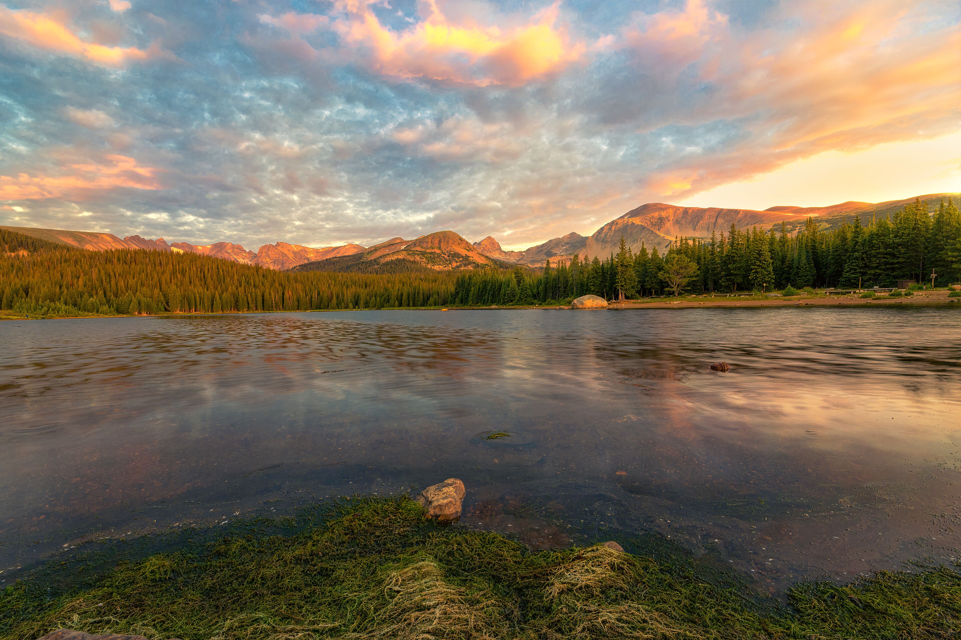 Un lago tranquilo rodeado de bosques densos y montañas bajo un cielo colorido.
