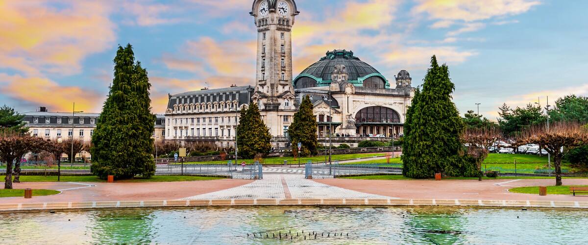 Un bâtiment historique doté d’une tour et d’un dôme imposants, entouré d’un parc bien entretenu avec une fontaine centrale.