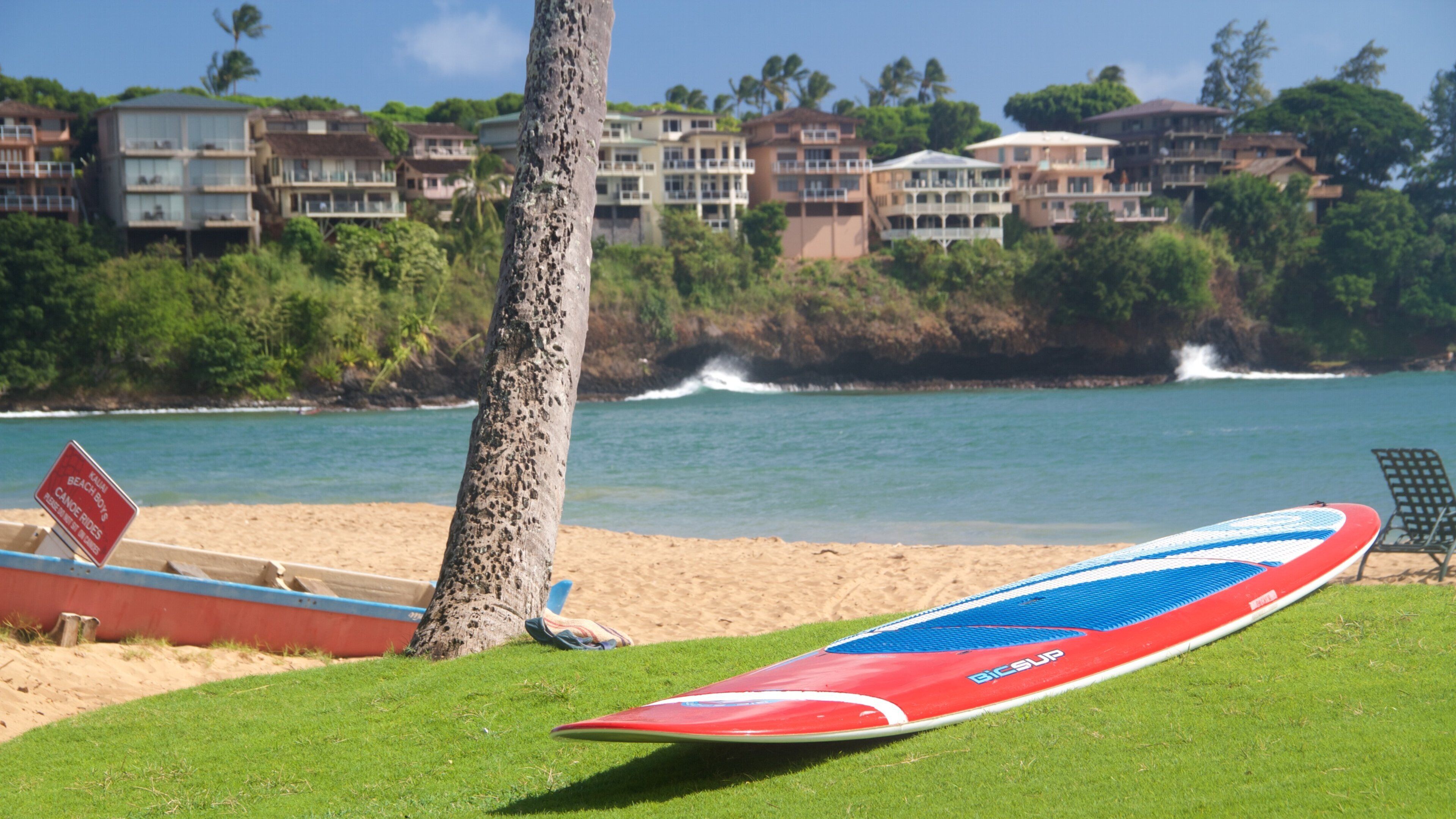 A beach with a surfboard, a sign for 'Kahului Beach Boys', and houses in the background.