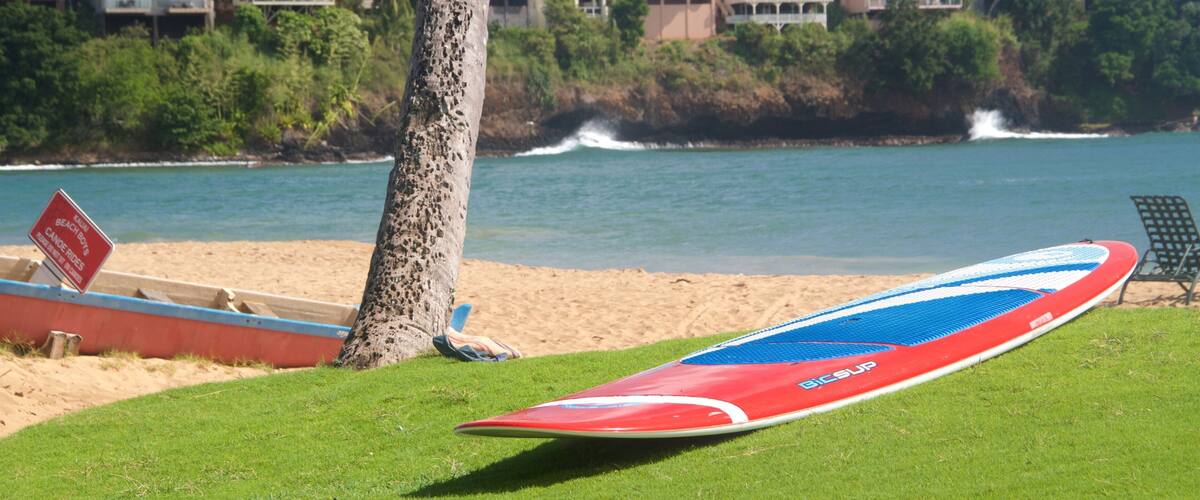 A beach with a surfboard, a sign for 'Kahului Beach Boys', and houses in the background.