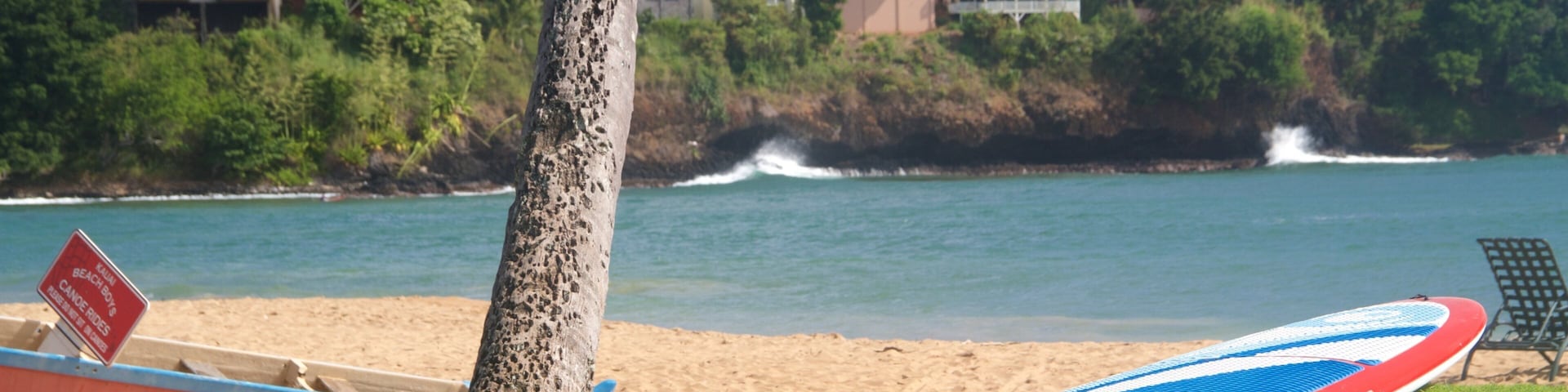 A beach with a surfboard, a sign for 'Kahului Beach Boys', and houses in the background.