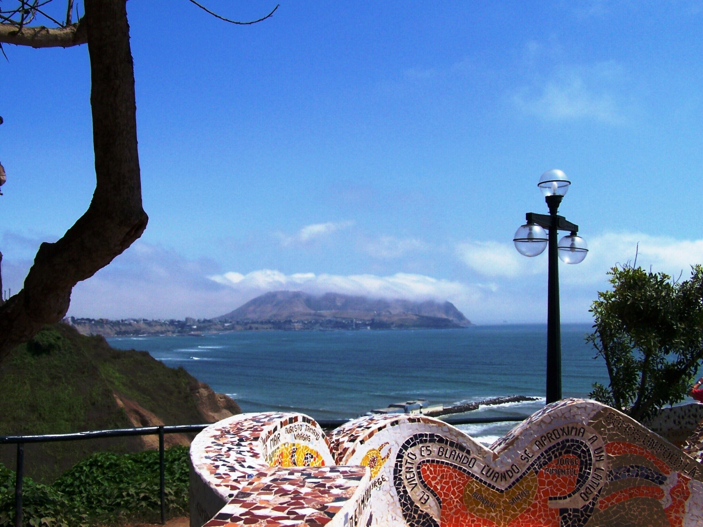 A coastal view with a mosaic bench, a lamp post, and a mountain in the distance.
