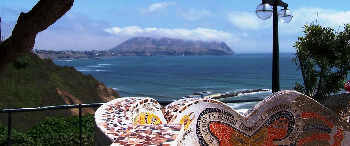 A coastal view with a mosaic bench, a lamp post, and a mountain in the distance.