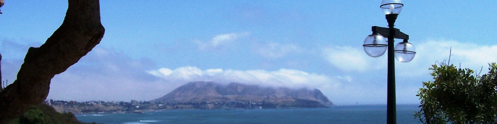 A coastal view with a mosaic bench, a lamp post, and a mountain in the distance.