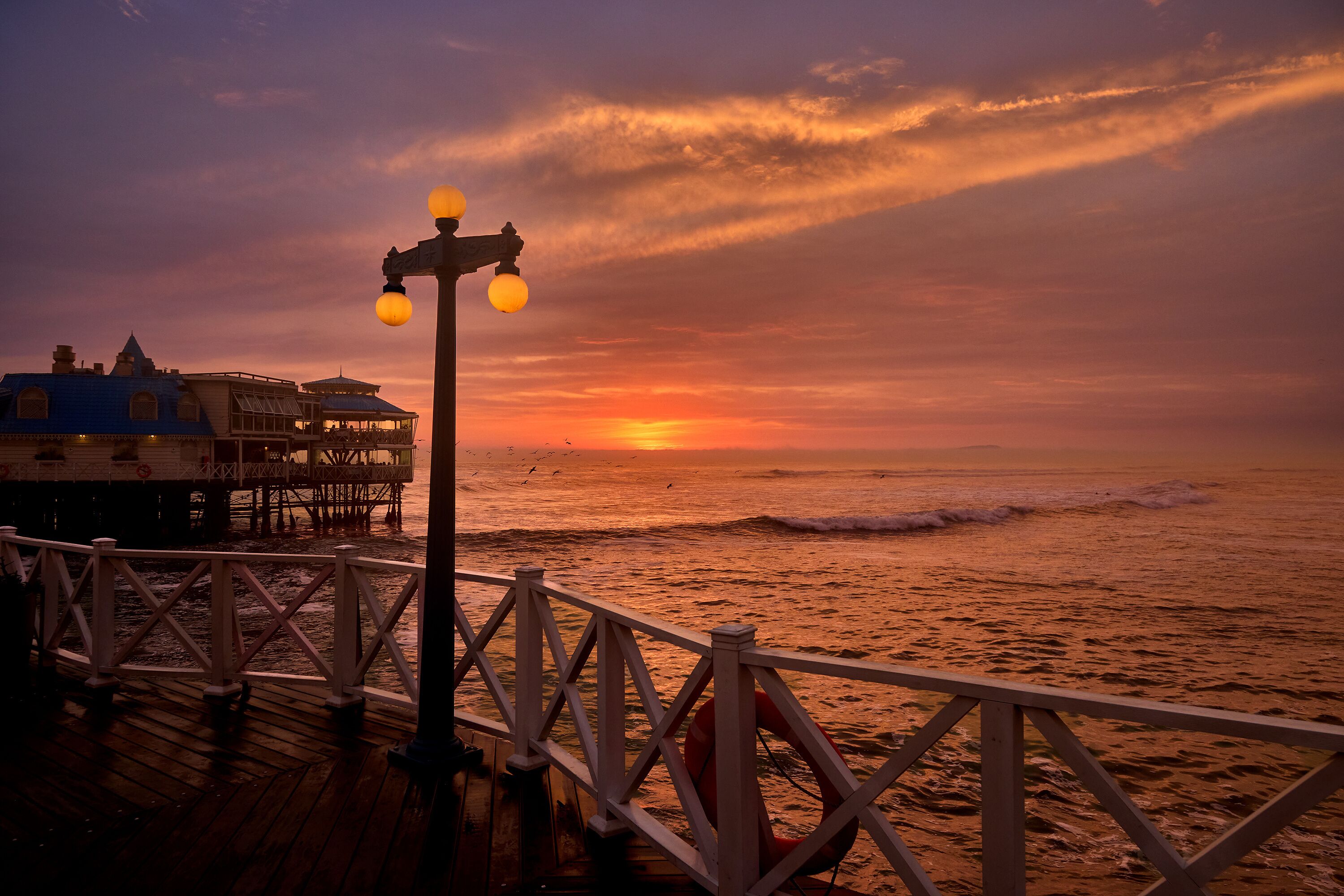 Una puesta de sol sobre el océano, con un muelle y edificios al fondo.