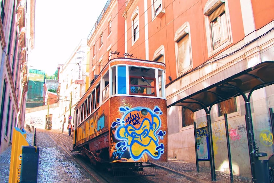 A colorful tram with graffiti on its side, traveling through a narrow street with buildings on either side.