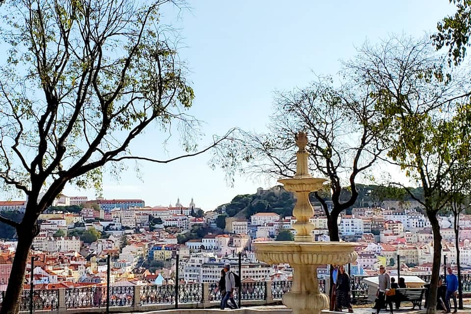 Ein Brunnen, umgeben von Bäumen, mit einer Stadtlandschaft im Hintergrund.